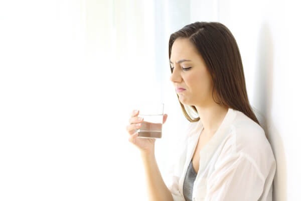 An image of a woman holding a water glass with a disgusted look on her face because her water smells like rotten eggs. 
